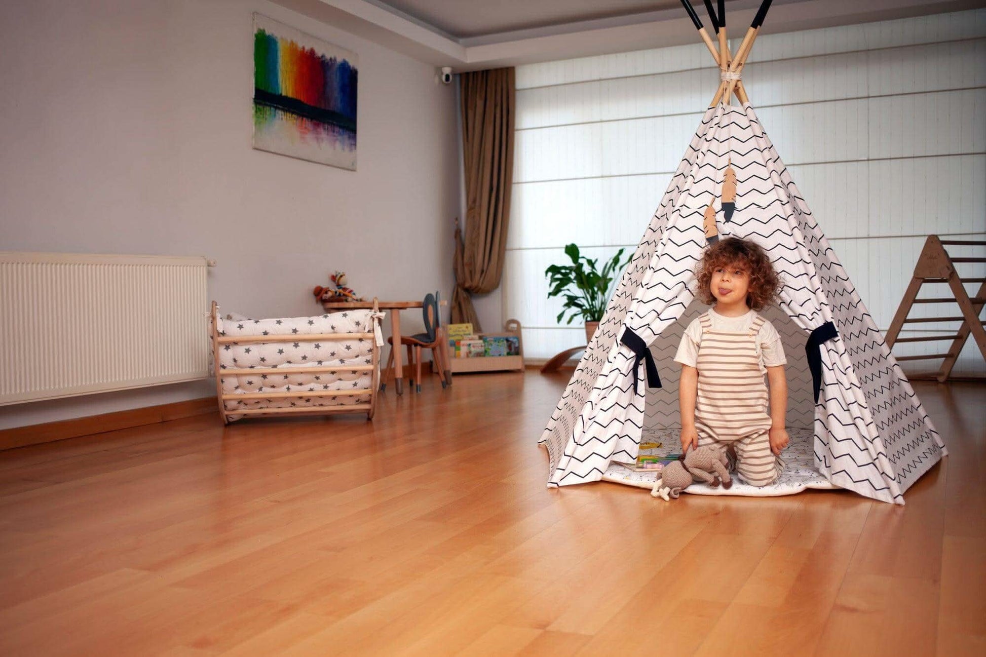 Child standing inside a teepee in a room with wooden floor and white walls.