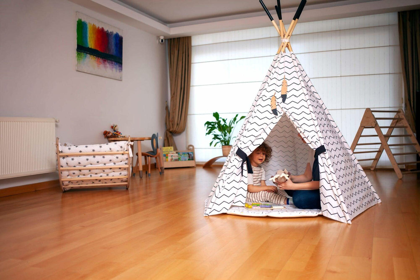 Children playing inside a teepee in a room with wooden floor and white walls.