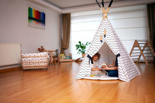 Children playing inside a teepee in a room with wooden floor and white walls.