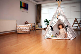 Child playing inside a teepee in a room with wooden floor and white walls.