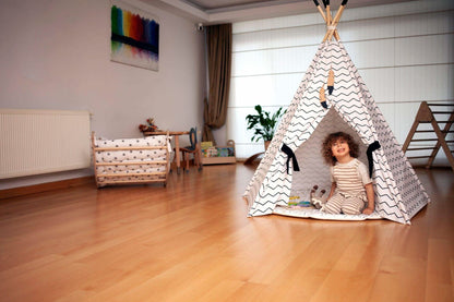 Child playing inside a teepee in a room with wooden floor and white walls.